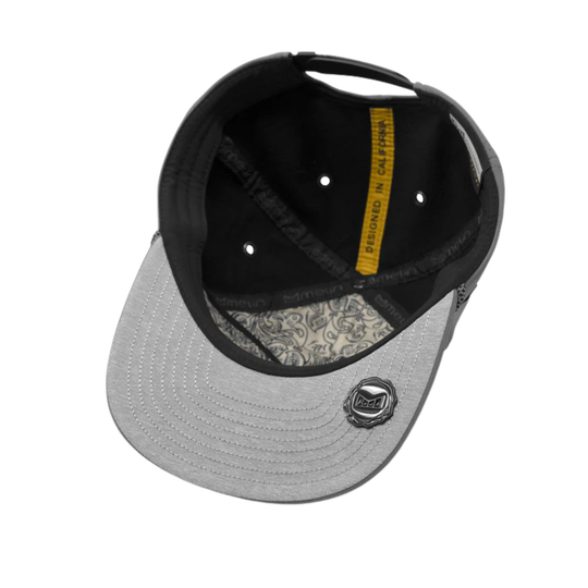 A structured baseball cap shown from a top-down angle, featuring a light gray curved brim, a black crown, a yellow vertical label reading 'DESIGNED IN california', and a small circular metal emblem on the brim.