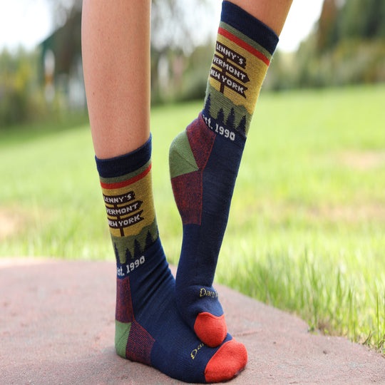 A close-up of a person's feet and lower legs wearing patterned crew socks outdoors on a stone path with a blurred green lawn in the background. The socks are navy blue with red, yellow, and green accents, featuring text that reads 'Lenny's Vermont NEW YORK' and 'est.