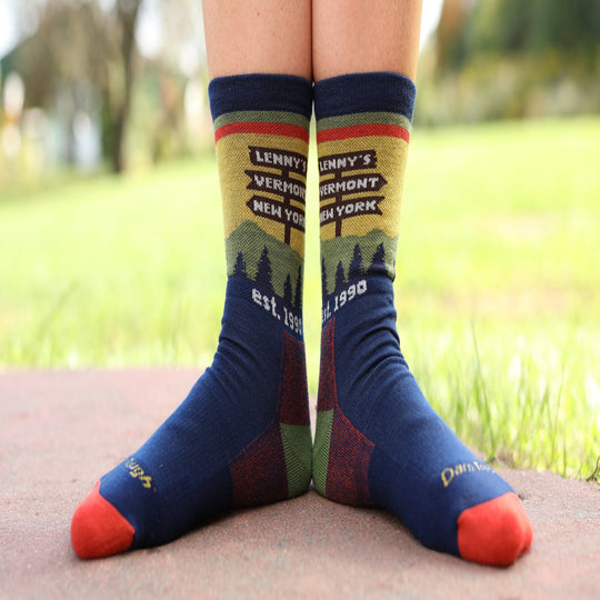 A person's feet wearing colorful crew socks with a scenic Vermont/New York graphic and the text 'Lenny's Vermont New York est. 1990' on a blurred park background.
