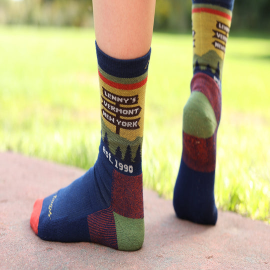 Close-up of a person's feet wearing colorful socks outdoors on a paved path with a blurred green lawn in the background. The socks feature a vintage-style travel graphic reading 'Lenny's Vermont New York Est.