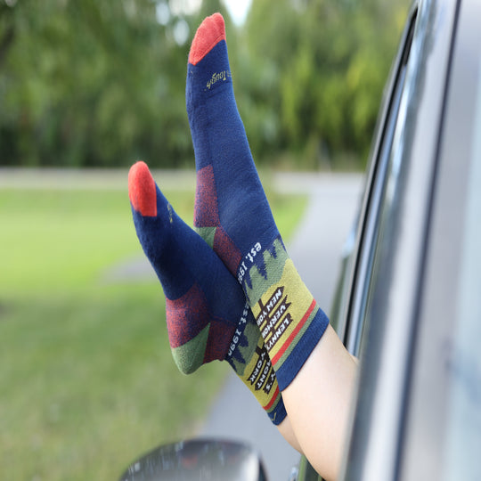 A person's legs sticking out of a car window wearing navy blue socks with red toes, heels, and cuffs, and a multicolor forest pattern on the soles. The socks have the brand name 'Lenny's' printed on them.
