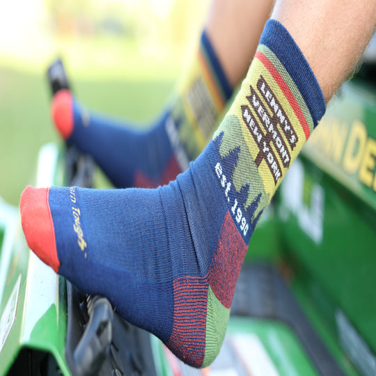 A close-up lifestyle shot of a person's feet wearing colorful crew socks while sitting in a green tractor. The socks are navy blue with red toes and heels, and feature a yellow, green, and red graphic design with the text 'LEWISY'S VERMONT NEW YORK est.