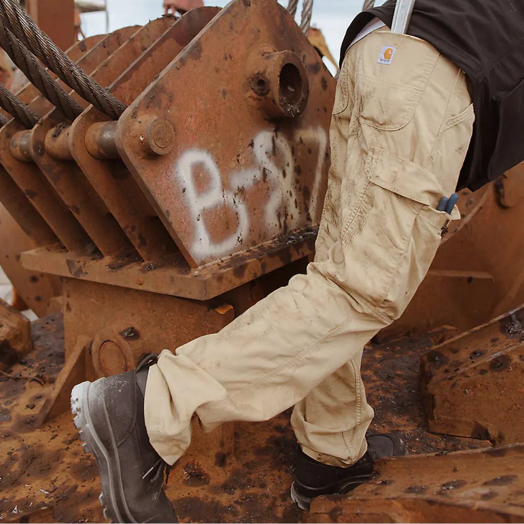 Person wearing beige work pants and black boots standing on a large rusty piece of machinery.