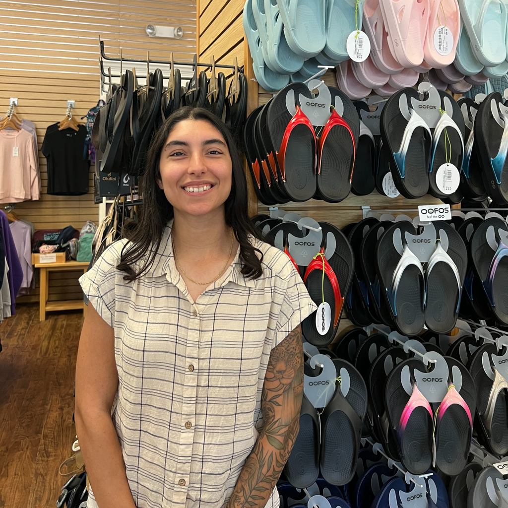 Woman standing in a store with Oofos sandals displayed on racks behind her.