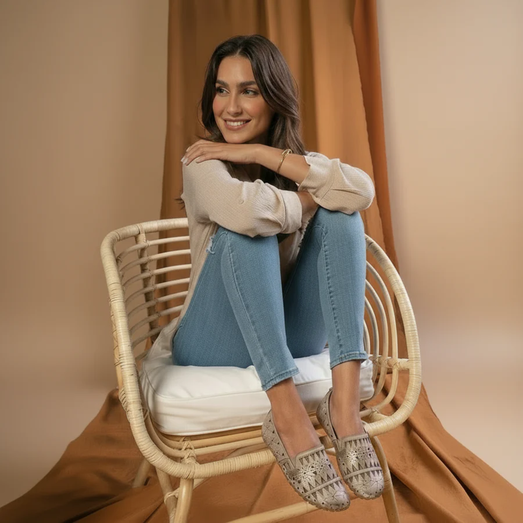 Woman sitting on a wicker chair wearing a beige sweater, blue jeans, and sandals with a neutral background.