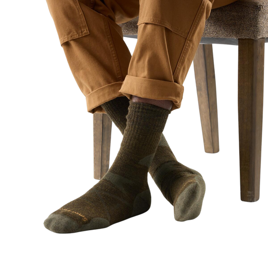 Person wearing olive socks and pants sitting on a wooden stool with a white background