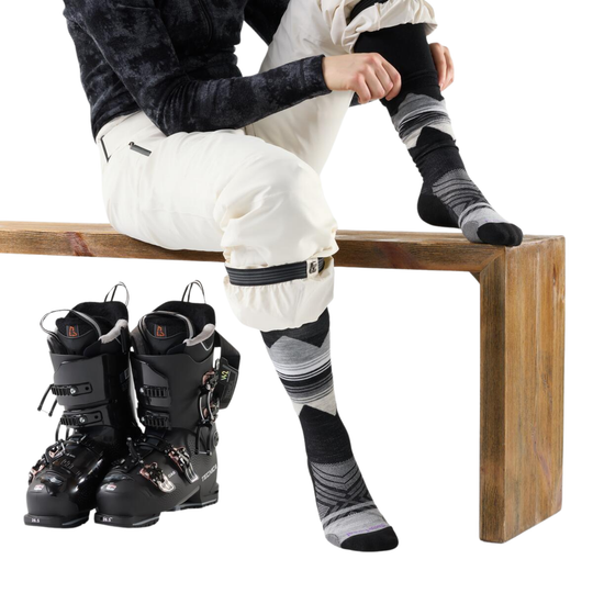 Person sitting on a wooden bench wearing ski boots and striped socks, with another pair of ski boots in front.