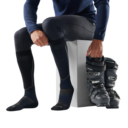 Person sitting on a white block wearing black ski boots and socks, with a white background.