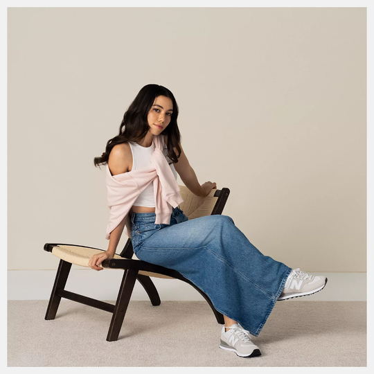 Woman sitting on a wooden bench wearing a white top, pink cardigan, blue jeans, and white sneakers.