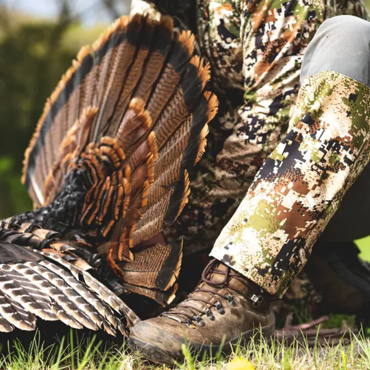 Person in camouflage clothing and boots interacting with a turkey in a natural setting