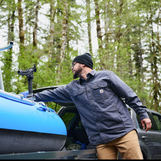 A man wearing a dark blue Carhartt work shirt, tan pants, and a black beanie leans against a blue inflatable raft on the back of a truck in a forest setting.