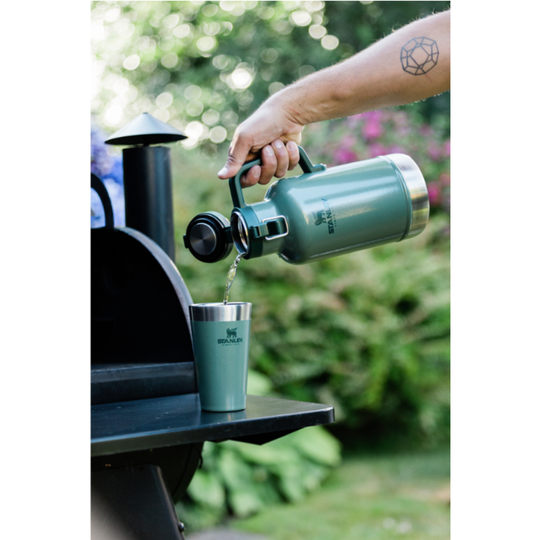 A person pours hot water from a matte sage green insulated kettle into a matching sage green travel tumbler, set against a blurred outdoor garden background.