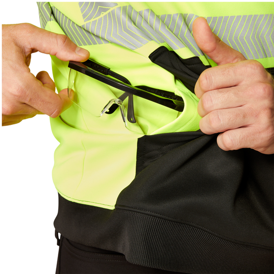 Close-up of a person wearing a high-visibility neon yellow-green jacket with reflective gray stripes, black lower paneling, and a side pocket holding black-framed glasses.