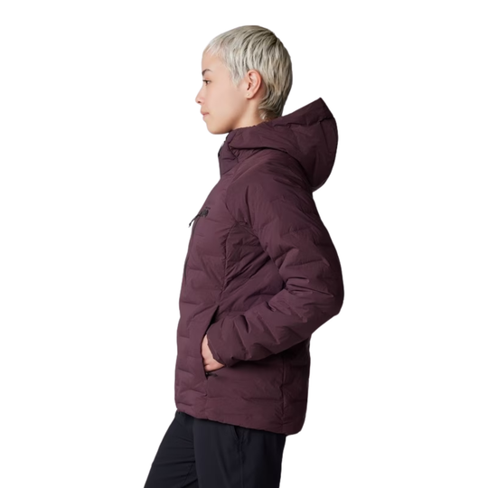 A side-profile view of a person wearing a hooded puffer jacket in a deep plum-burgundy color, paired with dark pants, against a plain white background.