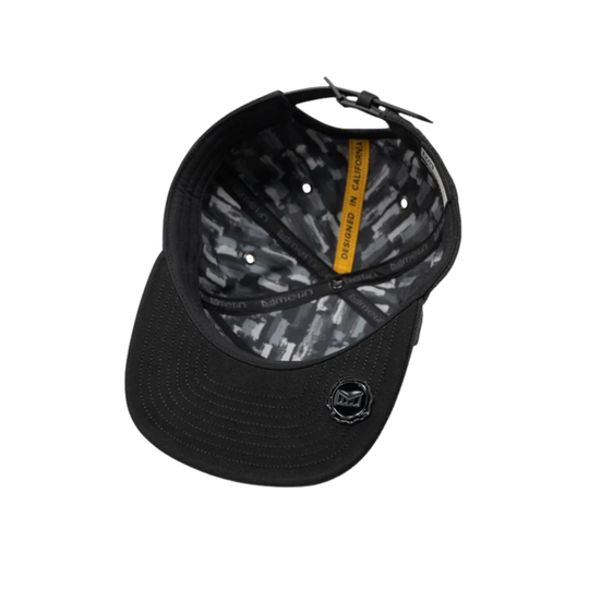 Black baseball cap with a black and gray geometric patterned interior, featuring a small black shield logo on the front of the brim and a yellow interior tag.