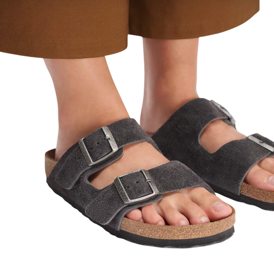 A close-up of a person wearing dark gray double-strap slide sandals with silver buckles and a cork-look footbed, shown against a white background with brown pants visible at the top.