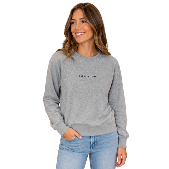 A woman wearing a heather gray crewneck sweatshirt with 'LIFE IS GOOD' printed in black across the chest, paired with light blue jeans, set against a plain white background.