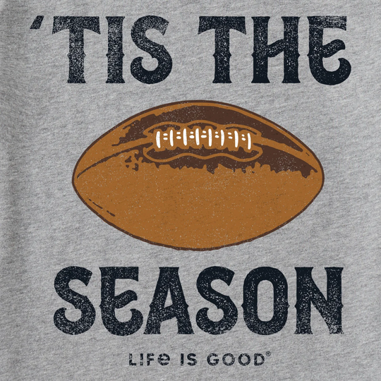 A close-up of a heather gray t-shirt featuring a distressed graphic print. The design displays the phrase "'TIS THE SEASON" in a dark navy blue vintage font, with a central brown football graphic and the text "LIFE IS GOOD" printed below it.
