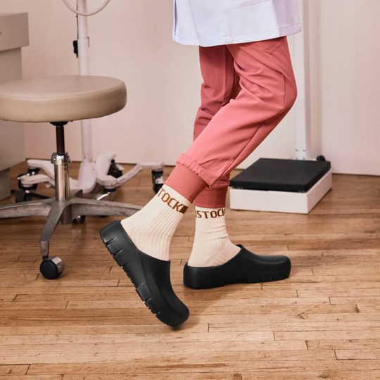 A person wearing black slip-on clogs and cream socks with gold 'BIRKENSTOCK' lettering, shown from the waist down in a clinical room with a wooden floor, stool, and medical equipment.