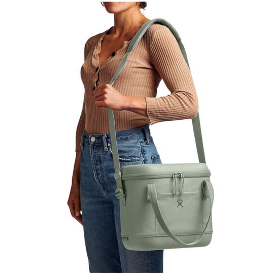 A woman wearing a beige ribbed long-sleeve top and blue jeans carries a sage green insulated cooler bag with a shoulder strap against a white background.