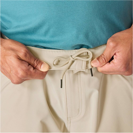 Close-up of a person wearing light beige drawstring shorts and a teal shirt, adjusting the waistband with both hands.