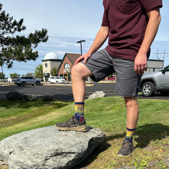 A person standing outdoors on a large gray rock, wearing dark brown GORE-TEX hiking shoes and colorful crew socks with a scenic mountain landscape print The background shows a grassy park with a Lenny's Shoe and Apparel store parking lot and brick buildings.