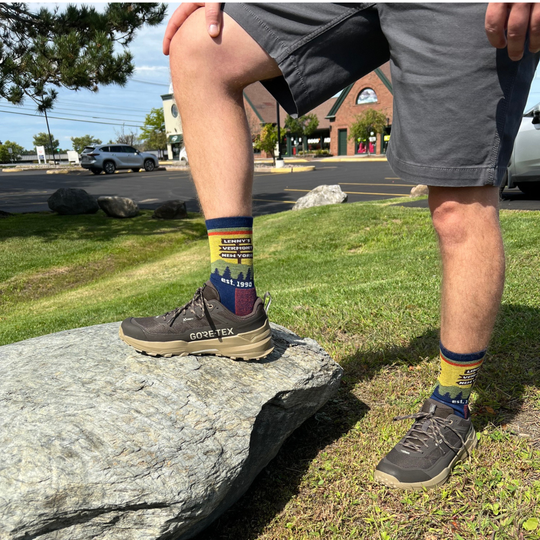A person standing outdoors on a large gray rock, wearing dark brown GORE-TEX hiking shoes and colorful crew socks with a scenic mountain landscape print The background shows a grassy park with a Lenny's Shoe and Apparel store parking lot and brick buildings.
