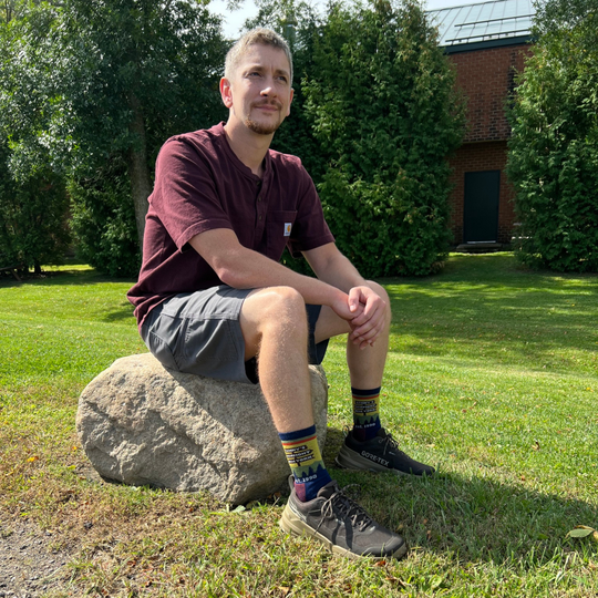 A person standing outdoors on a large gray rock, wearing dark brown GORE-TEX hiking shoes and colorful crew socks with a scenic mountain landscape print The background shows a grassy park with a Lenny's Shoe and Apparel store parking lot and brick buildings.