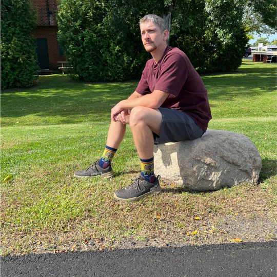 A person standing outdoors on a large gray rock, wearing dark brown GORE-TEX hiking shoes and colorful crew socks with a scenic mountain landscape print The background shows a grassy park with a Lenny's Shoe and Apparel store parking lot and brick buildings.