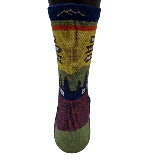 A single crew sock with a multicolor mountain landscape knit pattern, featuring a yellow and green hillside, dark blue sky and trees, and a red section near the foot, shown on a black mannequin leg against a white background.
