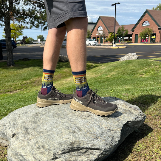 A person standing outdoors on a large gray rock, wearing dark brown GORE-TEX hiking shoes and colorful crew socks with a scenic mountain landscape print The background shows a grassy park with a Lenny's Shoe and Apparel store parking lot and brick buildings.