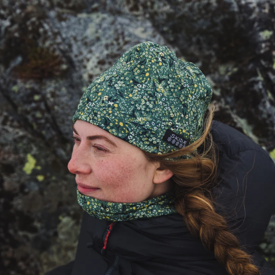 A woman wearing a green floral patterned beanie and matching neck gaiter, photographed outdoors against a rocky, mossy background. A small black brand tag on the beanie reads 'skida'.