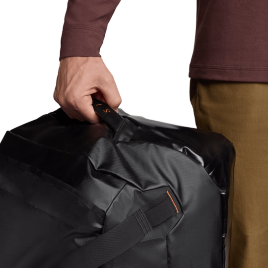 Person holding a black duffel bag with a strap, wearing a brown shirt and beige pants.