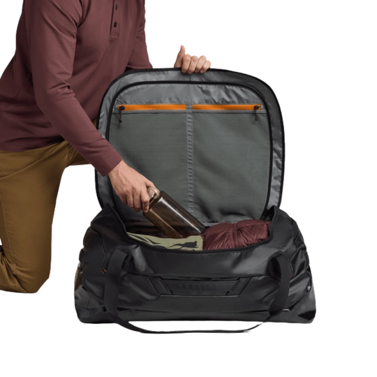 Person packing a large black duffel bag with items on a white background
