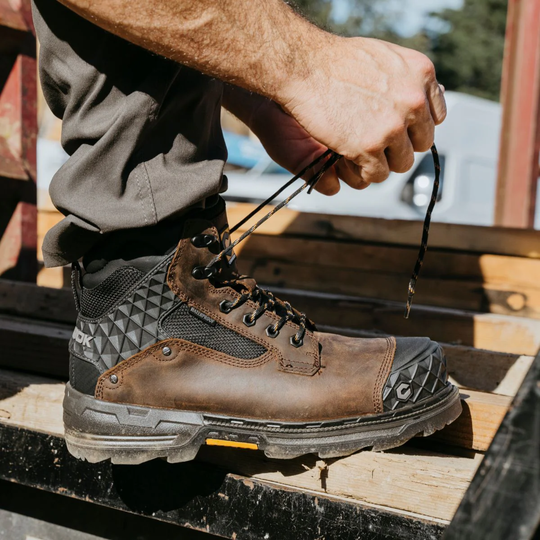 Person tying the laces of a brown and black work boot on a wooden surface.