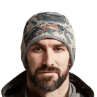 Person wearing a camouflage beanie with a brand logo on a white background