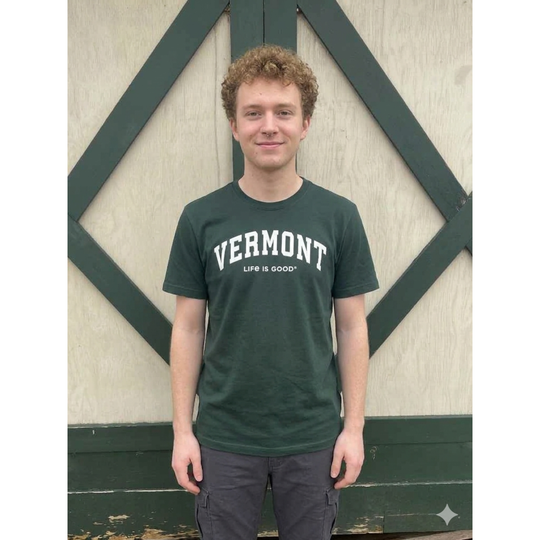 Person wearing a green 'Vermont Life is Good' t-shirt standing against a wooden background