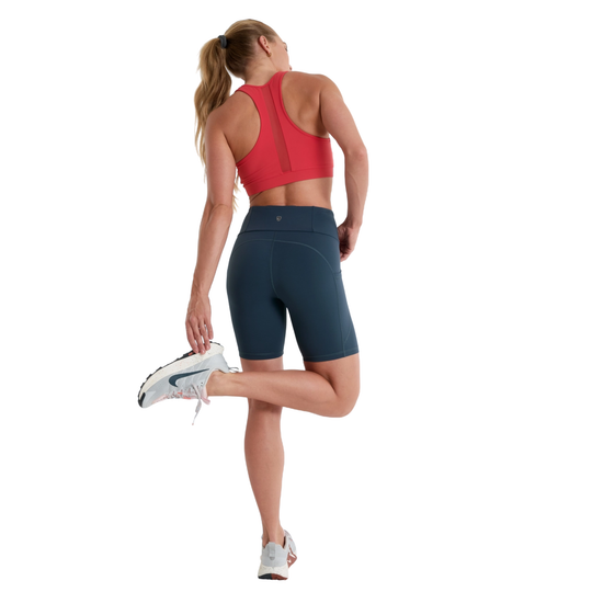 Woman in red sports bra and navy shorts stretching on a white background