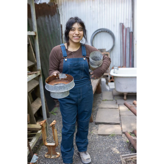 Woman wearing blue denim overalls and brown long-sleeve shirt, holding metal containers in a workshop yard
