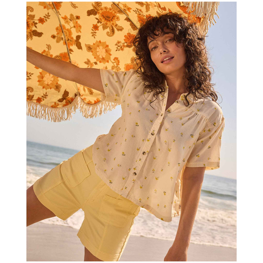 Woman on a beach holding a floral umbrella, wearing a light blouse and yellow shorts.