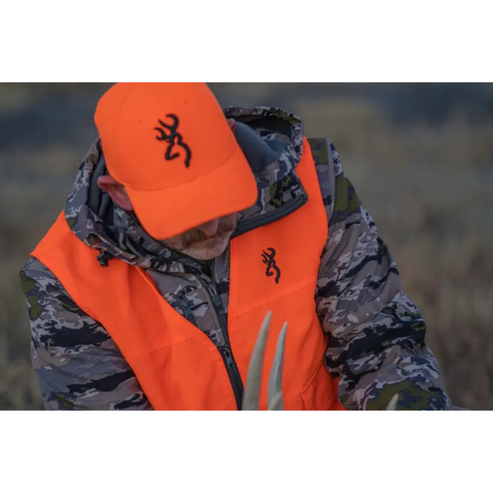Person wearing an orange hunting vest and cap with a logo, standing in a natural setting.