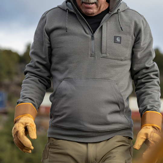 Man wearing a gray hoodie with a brand logo, tan gloves, and brown pants outdoors.