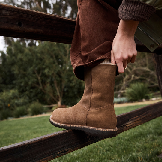 Person wearing brown boots and pants sitting on a wooden fence with a blurred natural background