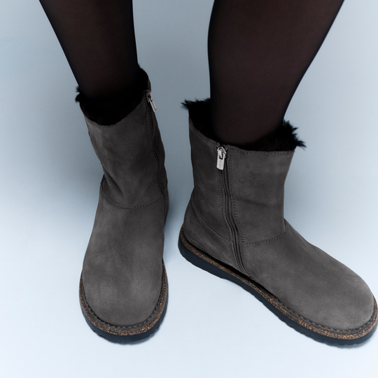 Gray suede boots with fur trim worn by a person on a light blue background
