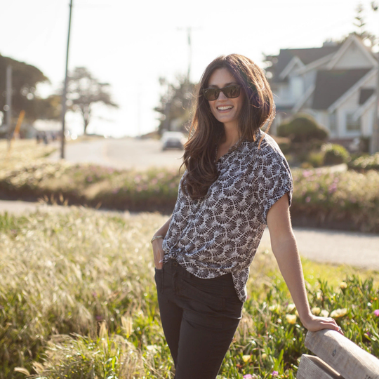 Woman standing outdoors in a residential area wearing sunglasses and a patterned top.