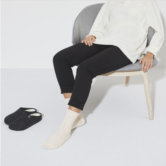 Person sitting on a chair wearing black pants, beige socks, and a white sweater with a pair of black slippers on the floor.