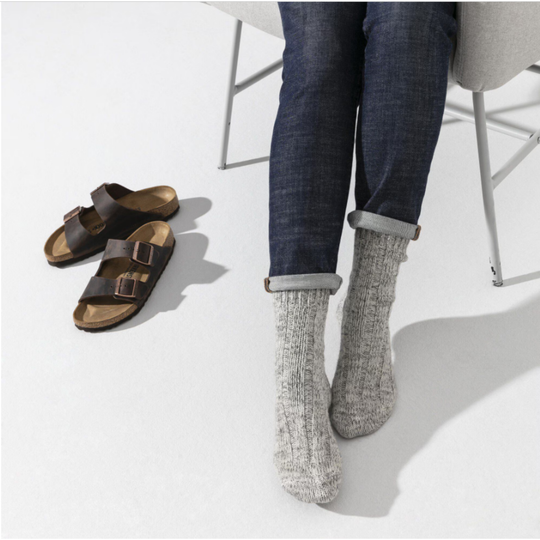 Person wearing gray socks and dark jeans with a pair of brown sandals on a white floor.