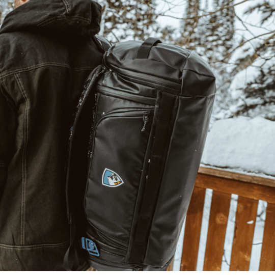 Person wearing a black jacket with a black backpack featuring a logo, standing outdoors in a snowy landscape.