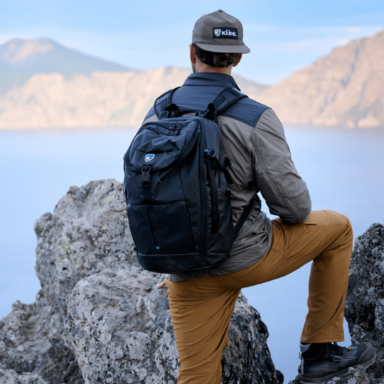 Person with a backpack sitting on a rock with mountains and water in the background