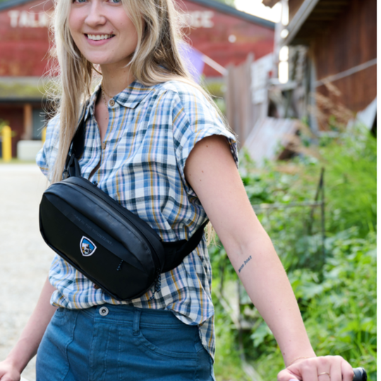 Woman wearing a black fanny pack with a logo, standing outdoors.